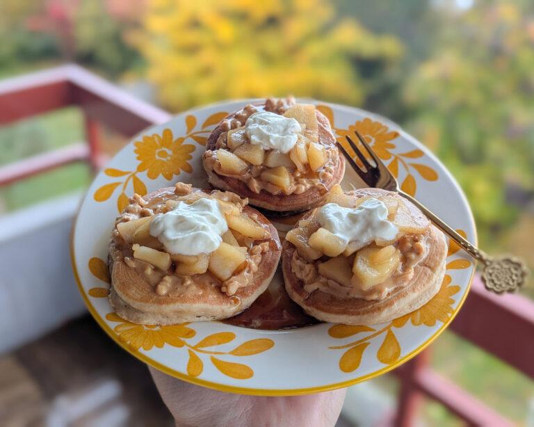 Assiette de pancakes moelleux garnis de pommes fondantes et d’un nuage de yaourt, servie sur un balcon avec vue automnale.