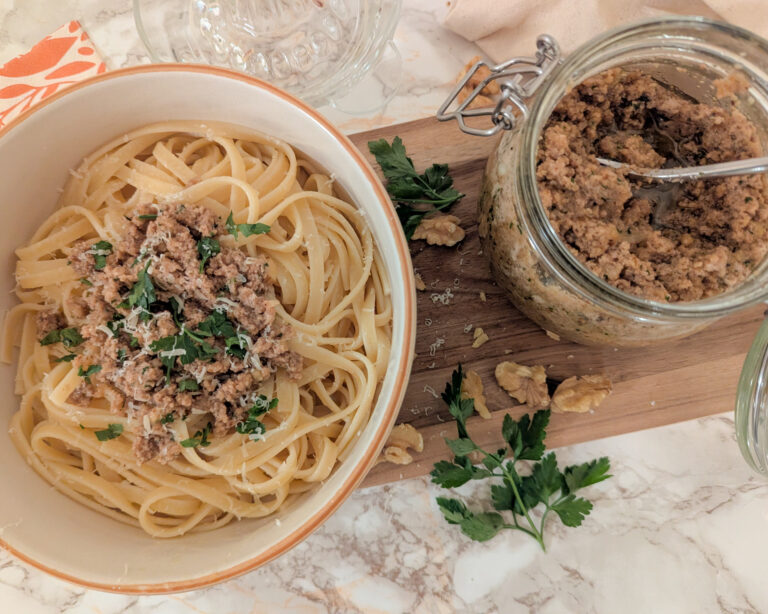 Assiette de spaghettis nappées d’un pesto aux noix maison, parsemées de persil frais, accompagnée d’un pot de pesto sur une planche en bois.