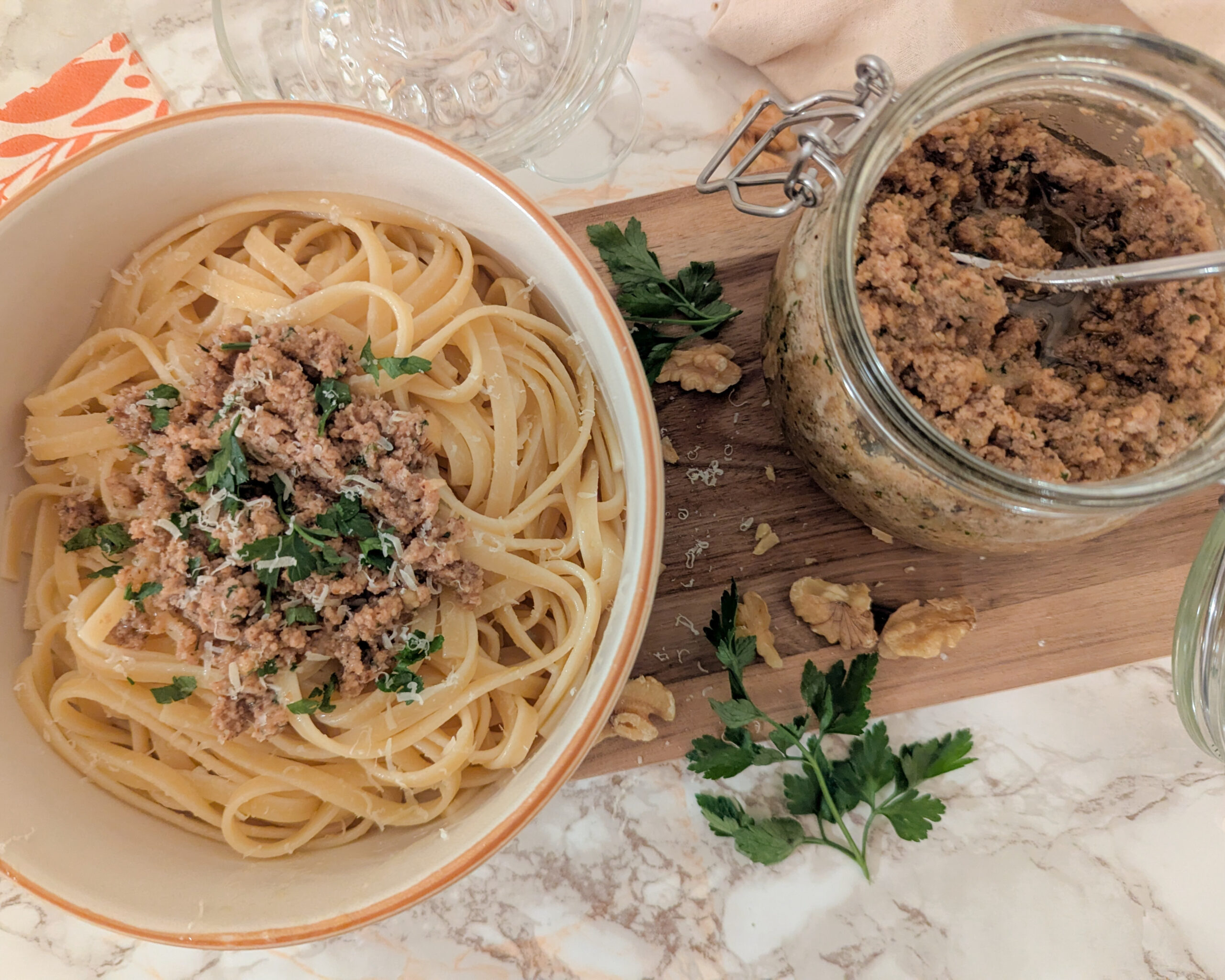Assiette de spaghettis nappées d’un pesto aux noix maison, parsemées de persil frais, accompagnée d’un pot de pesto sur une planche en bois.