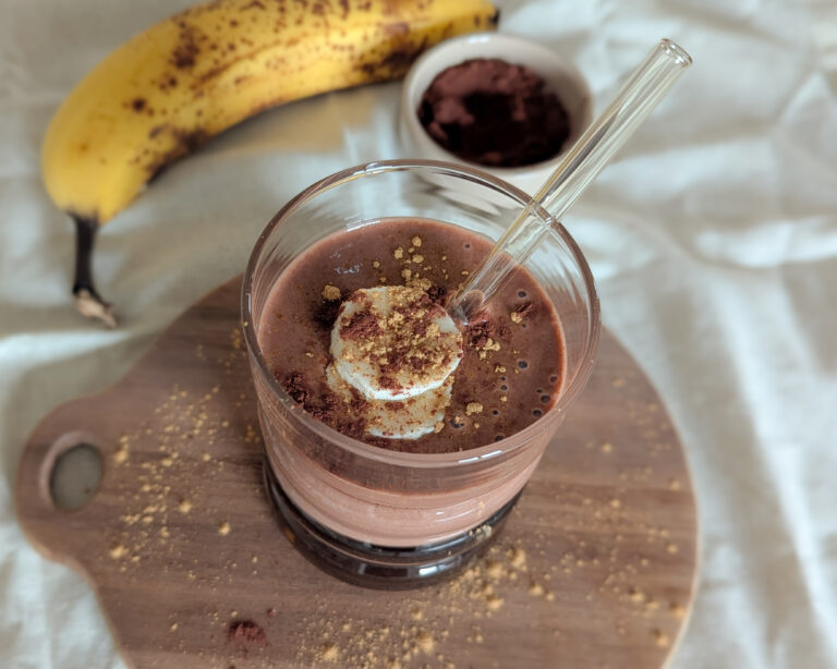 Verre de smoothie banane, cacao et cannelle, sur planche en bois avec une banane mûre et du cacao en poudre en arrière-plan.