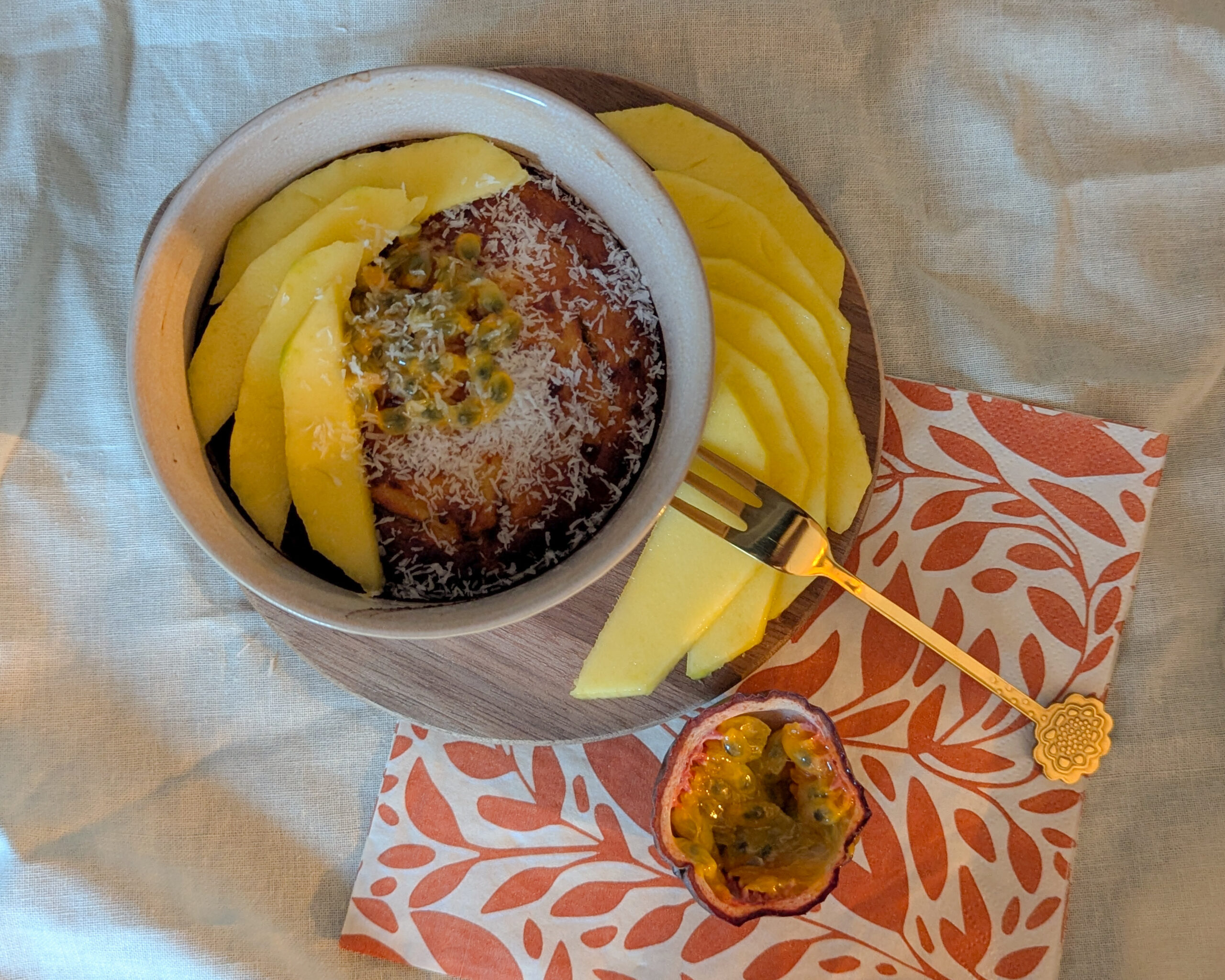 Bol de bowlcake à la farine de coco, accompagné de tranches de mangue fraîche et d’un demi-fruit de la passion, sur une serviette orange à motifs feuillus.