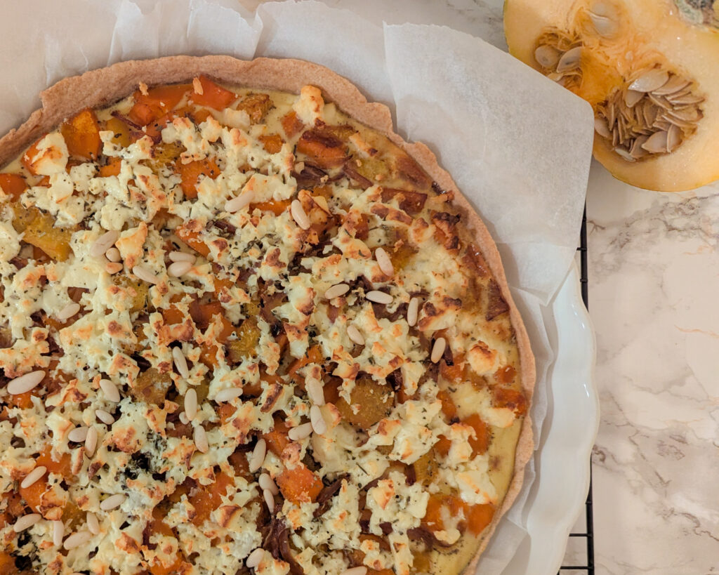Tarte maison à la courge butternut, feta et oignons rouges, parsemée de pignons de pin dorés, sur fond de linge crème.