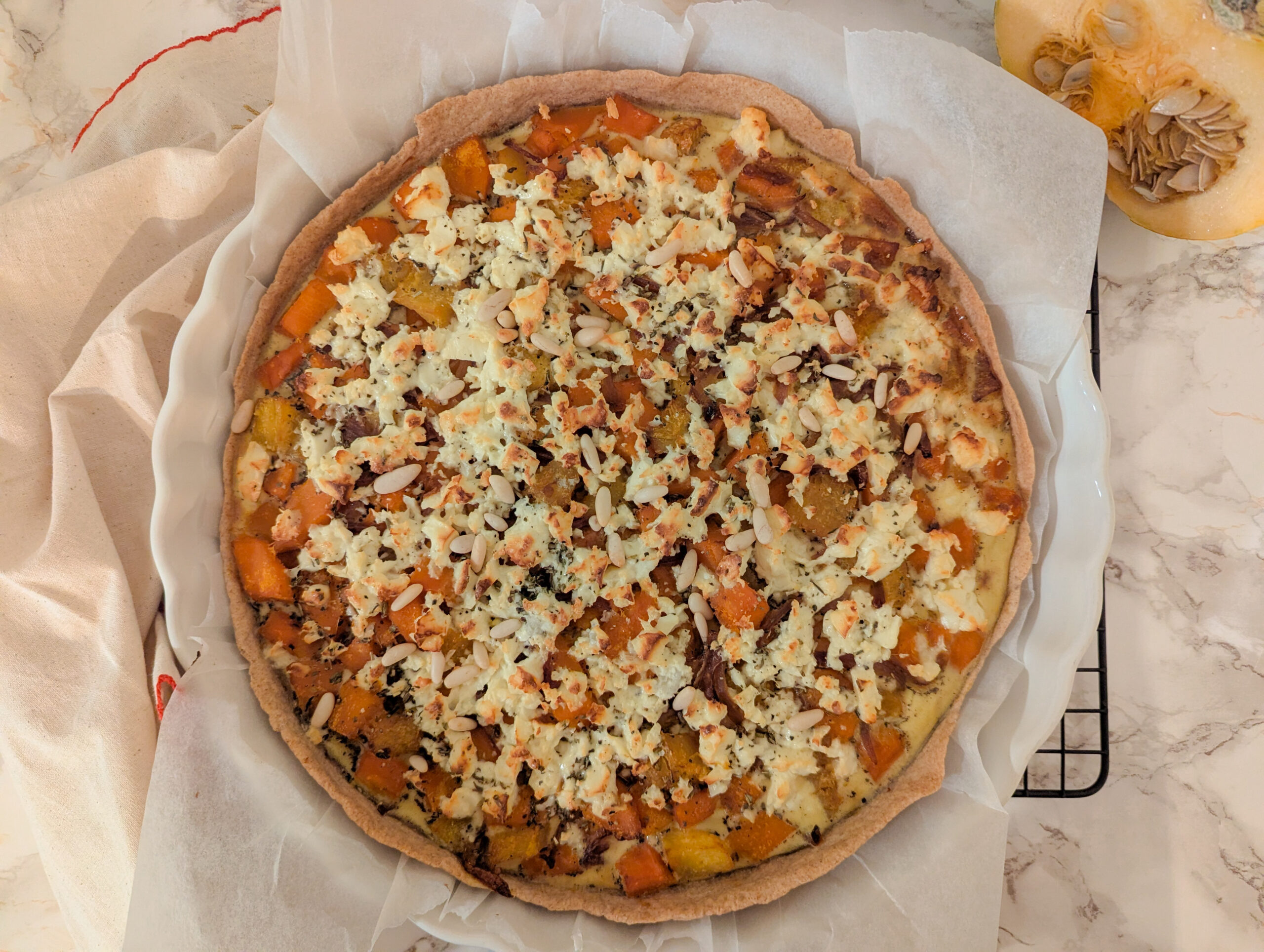 Tarte maison à la courge butternut, feta et oignons rouges, parsemée de pignons de pin dorés, sur fond de linge crème.