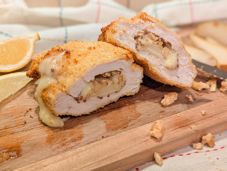 Cordon bleu de poulet croustillant garni de moutarde à l’ancienne, gruyère fondant, poire et éclats de noix, coupé en tranches sur une planche en bois.