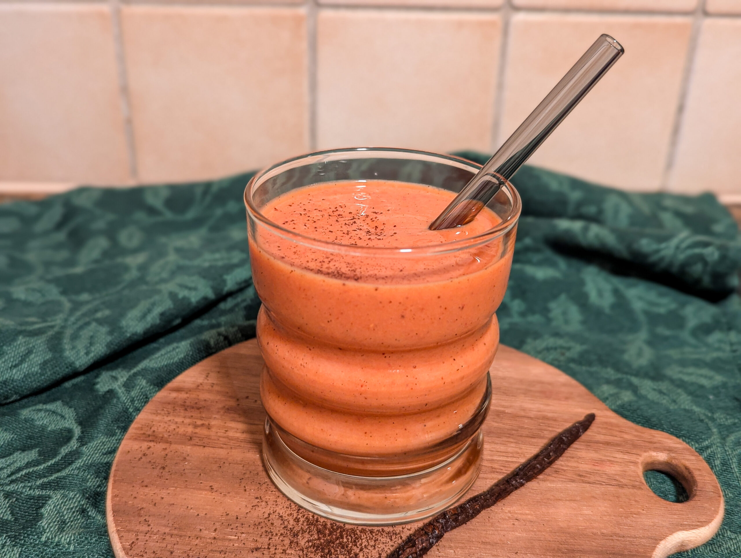 Smoothie orange dans un verre posé sur une planche en bois, avec une paille réutilisable, sur un fond vert.
