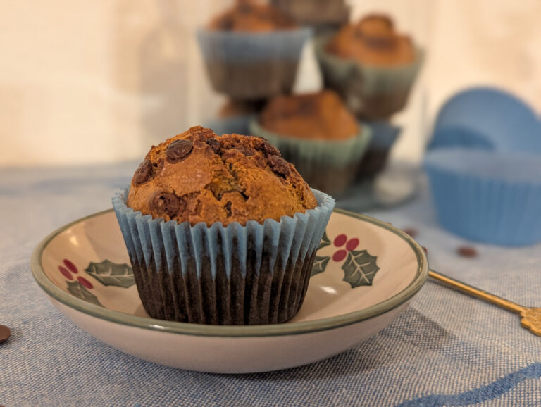 Un muffin banana bread doré et moelleux posé dans une caissette bleue, présenté dans une petite assiette vintage, avec d’autres muffins flous en arrière-plan.