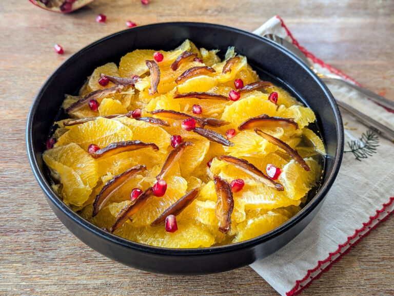 Salade d’oranges tranchées, dattes et graines de grenade nappées de sirop épicé, servie dans un bol noir sur une table en bois.