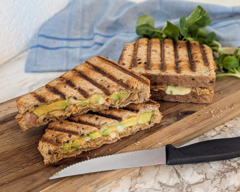 Croque-monsieurs grillés garnis de poulet effiloché, avocat et fromage, coupés en triangles et présentés sur une planche en bois avec un couteau.