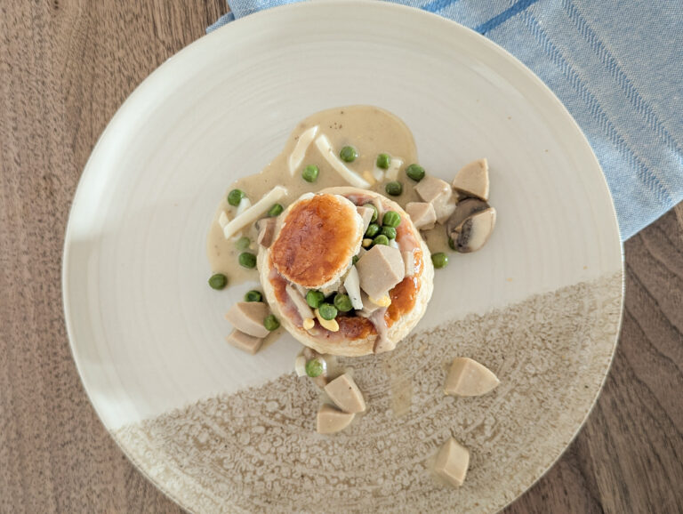 Vol-au-vent maison garni de sauce crémeuse aux champignons, petits pois et saucisses, servi dans une assiette claire vue de dessus.